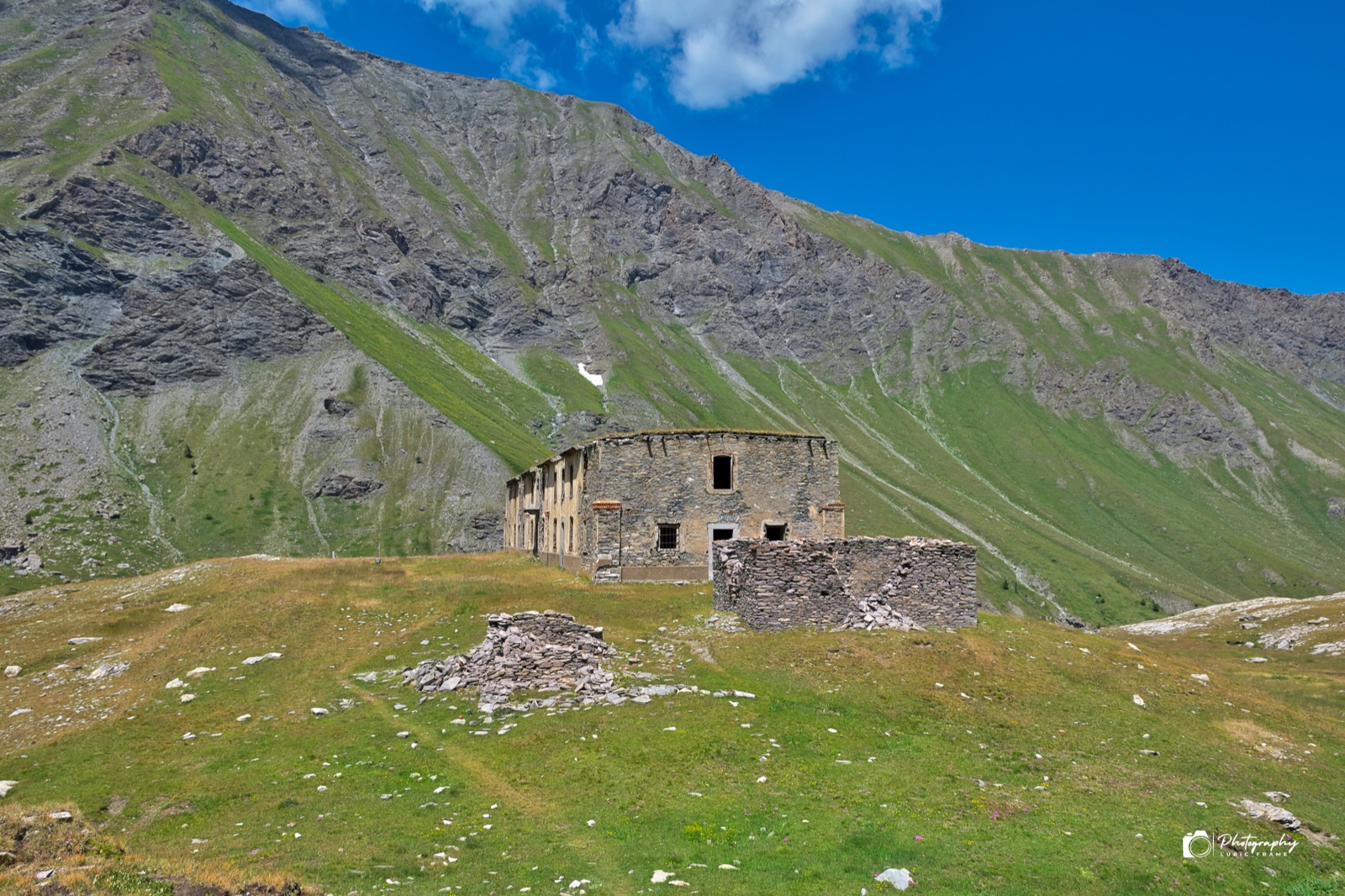 Rovine di un edificio in pietra su un pianoro erboso ai piedi di un pendio montuoso