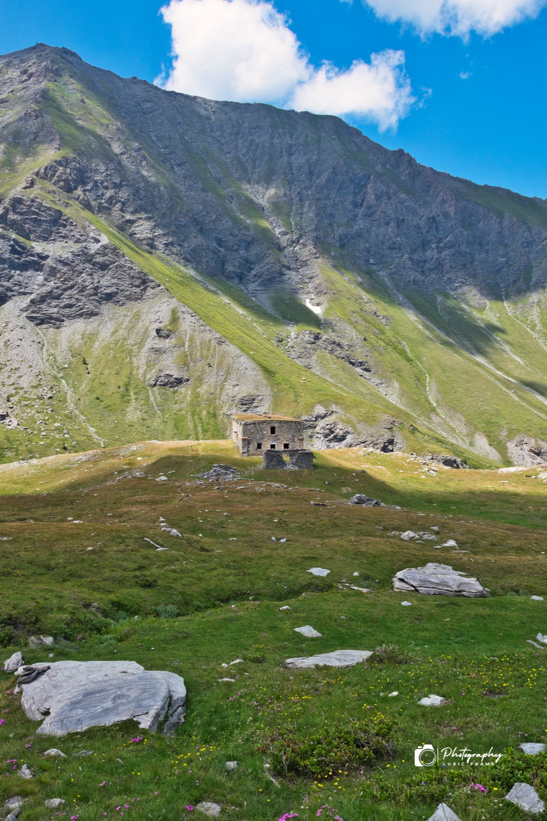 Vecchia costruzione in pietra in un pascolo d'alta quota, con un grande masso in primo piano e una montagna alle spalle