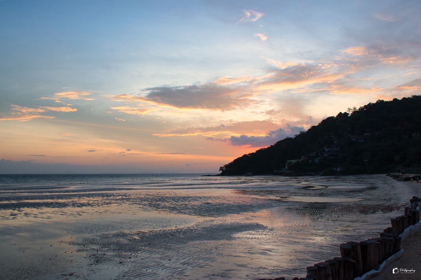 Spiaggia bagnata che riflette il cielo del tramonto con le nuvole, e un promontorio scuro sulla destra