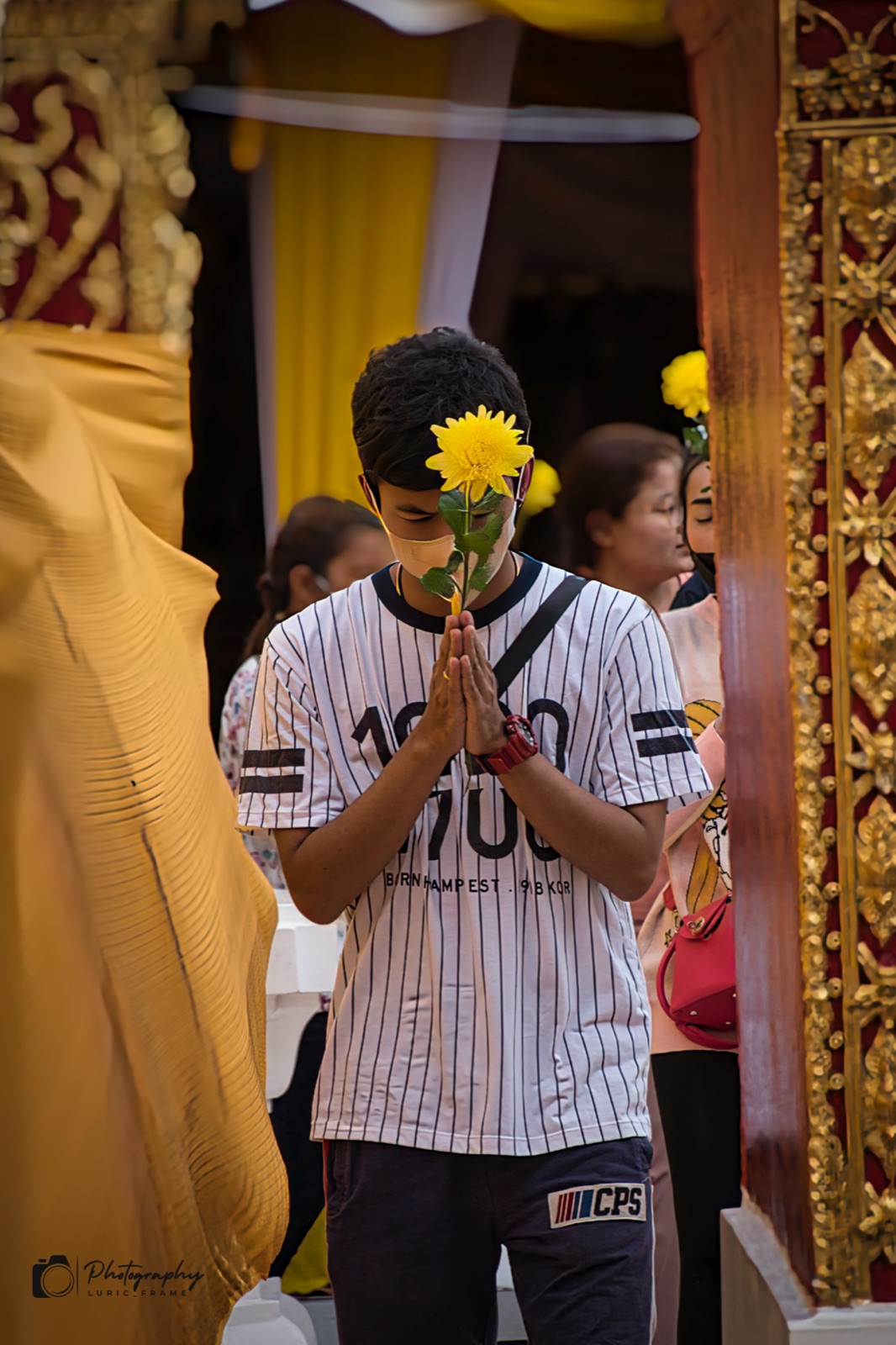 Ragazzo con le mani giunte che stringe un fiore giallo, in un tempio dalle decorazioni dorate