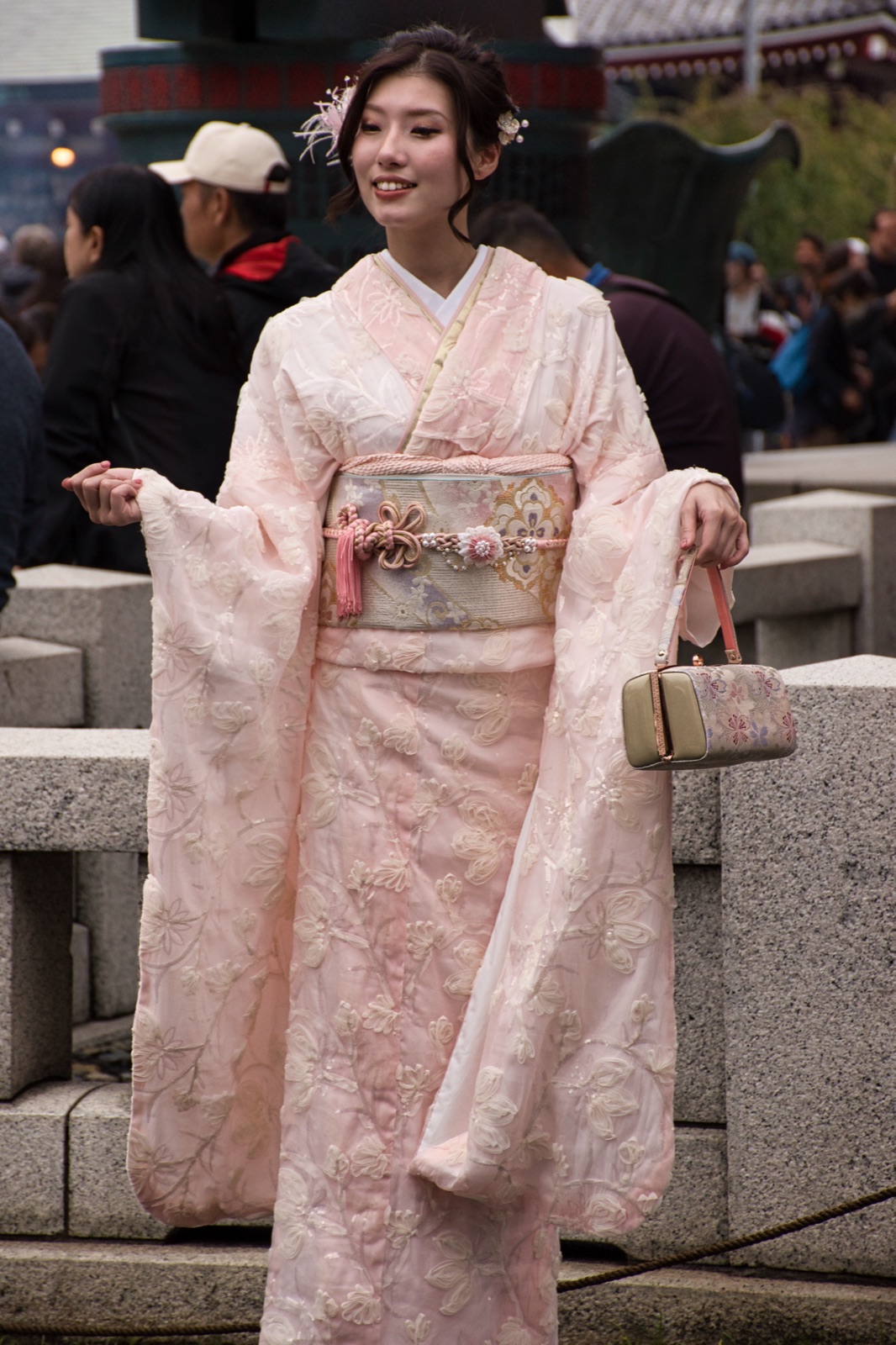 Ragazza in kimono rosa ritratta a figura intera su un piazzale in pietra, con una piccola borsa in mano