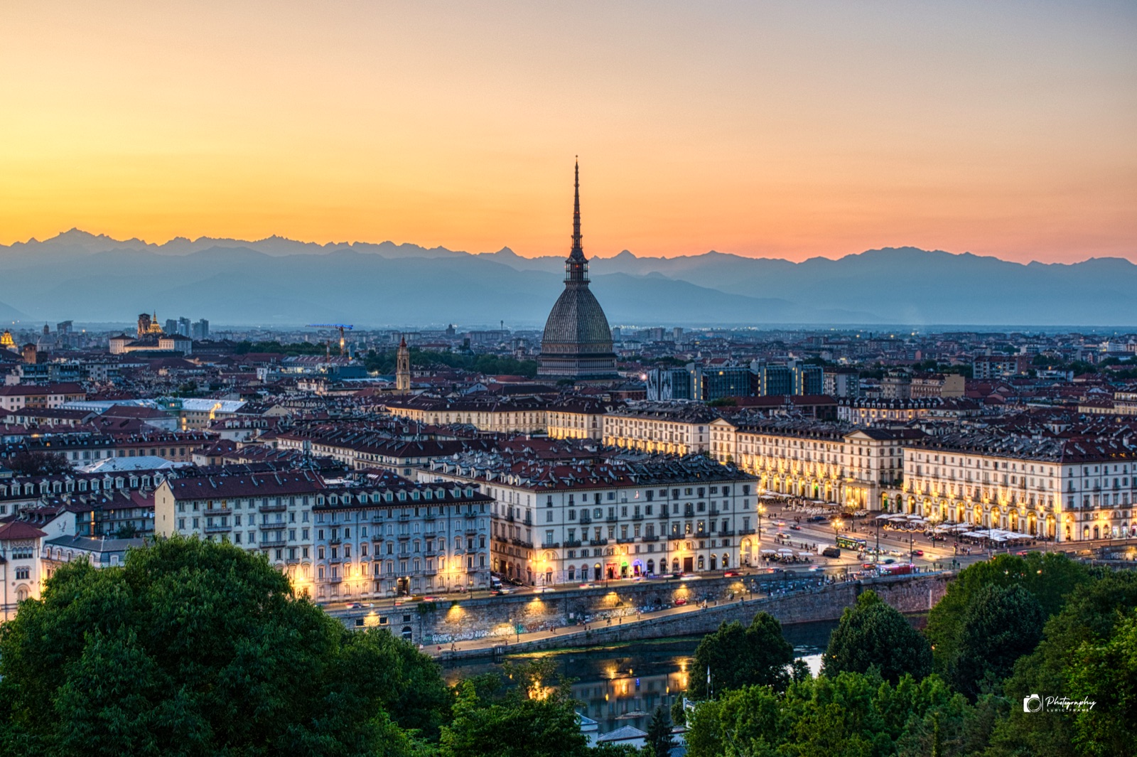 Città al tramonto vista dall'alto, con un'alta guglia al centro, le prime luci accese e le Alpi all'orizzonte